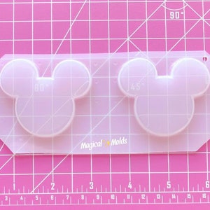 Puede incluir: Un molde de plástico transparente para hacer dos manualidades de resina con forma de Mickey Mouse. El molde está sobre una estera de corte rosa con líneas de cuadrícula blancas y el texto "Magical Molds" está impreso en el molde.