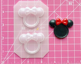 Puede incluir: Un molde de silicona rosa con dos diseños de anillos de Minnie Mouse y una cabeza de Minnie Mouse terminada. El molde tiene forma de lazo y anillo. La cabeza terminada es negra con un lazo rojo y orejas negras. El texto "Magical Molds" es visible.