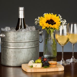 May include: A silver metal ice bucket with a bottle of white wine and ice inside. Two glasses of white wine are on a wooden cutting board with cheese, grapes, and berries.