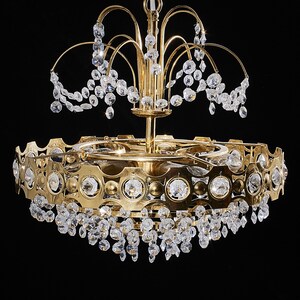 May include: A gold-toned chandelier with multiple tiers and crystal accents. The chandelier has a circular design with a series of clear crystals hanging from the bottom and top. The fixture is suspended from a chain against a black background.