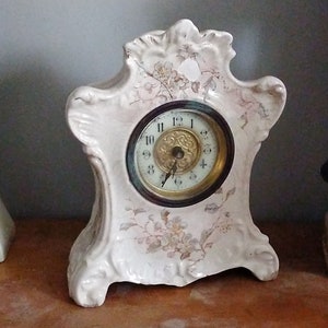 May include: A white porcelain mantel clock with floral details and a gold-toned face. The clock has a Roman numeral dial and a black hand.