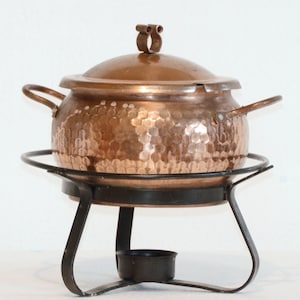 May include: A hammered copper fondue pot with a lid and two handles sits on a black metal stand with a candle holder.