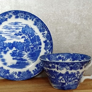May include: A blue and white porcelain teacup and saucer set with a floral pattern and a landscape scene. The saucer features a blue and white floral border with a central scene of a bridge over a river with a house and trees in the background. The teacup has a similar floral pattern and a landscape scene with a house and trees.