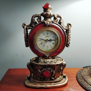 May include: A gold-toned decorative clock with a red face and Roman numeral markings. The clock has a detailed ornate design with a red jewel on the base.