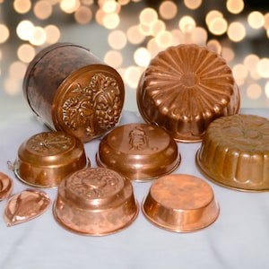 Może przedstawiać: Zbiór vintage form do ciasta z miedzi o różnych wzorach, w tym winorośl, osoba i wzór żebrowany. Formy są ułożone na białym tle.