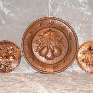 Könnte beinhalten: Drei dekorative Kupferplatten mit geprägten Obstmotiven. Die größte Platte zeigt Birnen, während die kleineren Teller Äpfel und Kirschen darstellen. Die Platten haben eine warme, rötlich-braune Farbe und sind wahrscheinlich Vintage.
