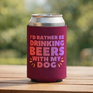 May include: A maroon beer can cooler with the text "I'D RATHER BE DRINKING BEERS WITH MY DOG" in a gradient of orange, pink, and purple. The cooler is on a wooden surface with a blurred green background.