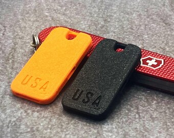 May include: An orange and a black phone case with the text "USA" are displayed next to a red Swiss Army knife. The knife has a white cross emblem and a small silver key ring.