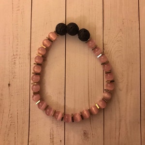 May include: A pink and black beaded bracelet with gold accents. The bracelet features pink and white beads with a gold trim, and three black lava rock beads.