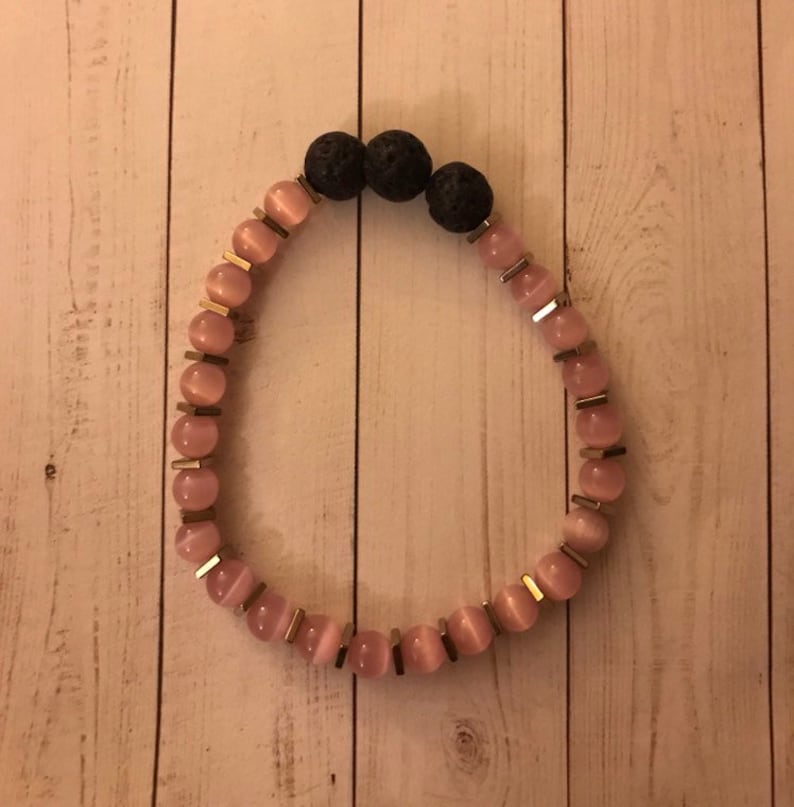 May include: A pink and black beaded bracelet with gold accents. The bracelet features pink and white beads with a gold trim, and three black lava rock beads.