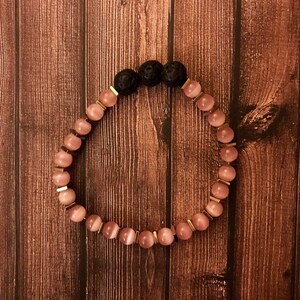 Puede incluir: Un brazalete de cuentas rosa y dorado con cuentas de lava negra. El brazalete está hecho con piedras naturales y está diseñado para ser usado en la muñeca.