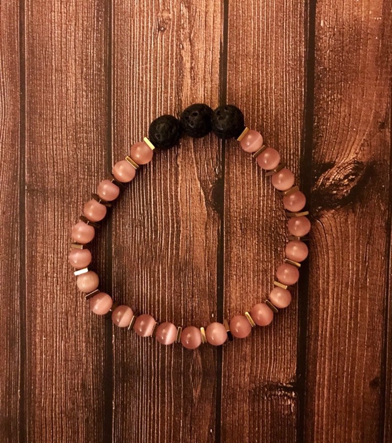 May include: A pink and gold beaded bracelet with black lava beads. The bracelet is made with natural stones and is designed to be worn on the wrist.