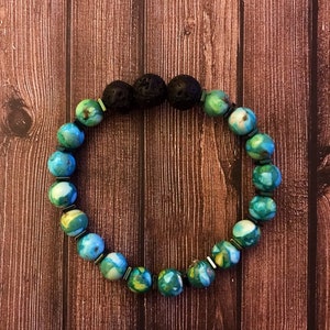 Puede incluir: Una pulsera de cuentas redondas grandes en tonos azules, verdes y blancos, intercaladas con pequeños espaciadores metálicos. La pulsera también incluye tres cuentas de lava negras y porosas. La pulsera se exhibe sobre una superficie de madera.