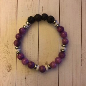 Puede incluir: Un brazalete de cuentas moradas y negras con detalles plateados. El brazalete está hecho con cuentas de lava y amatista.
