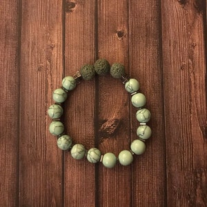 Puede incluir: Un brazalete de cuentas verdes y plateadas con cuentas de roca volcánica. El brazalete está sobre un fondo de madera marrón.