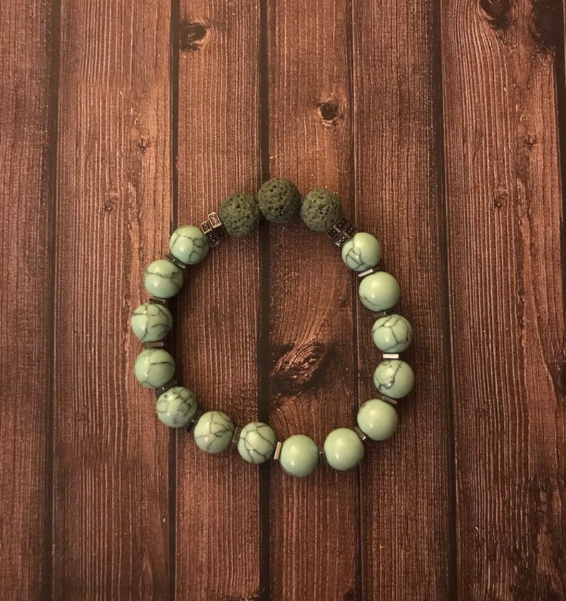 May include: A green and silver beaded bracelet with lava rock beads. The bracelet is on a brown wooden background.