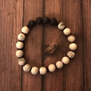 Puede incluir: Un brazalete hecho de cuentas blancas y negras. El brazalete tiene una mezcla de cuentas redondas y cuadradas. Las cuentas están enhebradas en un cordón marrón.