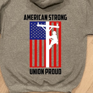 May include: A gray hooded sweatshirt with the text "AMERICAN STRONG" and "UNION PROUD" printed on the back. The design features a silhouette of a person climbing a utility pole with an American flag in the background.