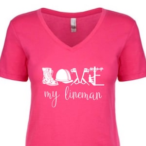 May include: Pink V-neck t-shirt with a white graphic design that says "Love my lineman" and includes illustrations of a hard hat, boots, and tools.
