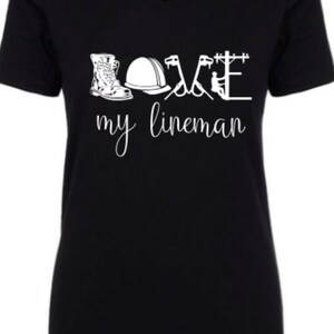 May include: Black v-neck t-shirt with a white graphic design that says "LOVE my lineman" and includes a boot, hard hat, pliers, and a lineman on a power pole.