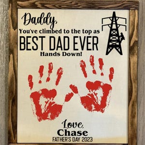 May include: A framed sign with a hand-painted message that reads "Daddy, You've climbed to the top as BEST DAD EVER Hands Down!" with two red handprints and the text "Love, Chase FATHER'S DAY 2023".