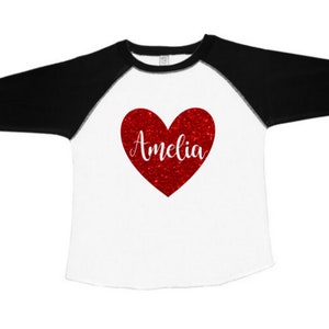 K&ouml;nnte beinhalten: Ein wei&szlig;es und schwarzes Baseball-T-Shirt mit einem roten Glitzerherz, auf dem "Amelia" steht.