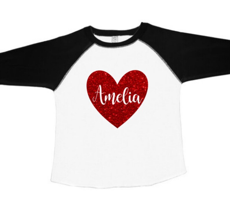 K&ouml;nnte beinhalten: Ein wei&szlig;es und schwarzes Baseball-T-Shirt mit einem roten Glitzerherz, auf dem "Amelia" steht.