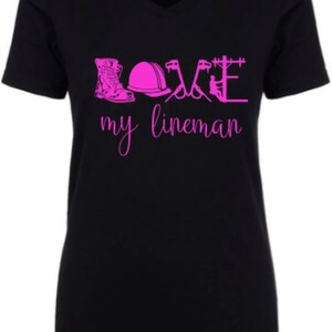 May include: Black v-neck t-shirt with pink lettering that reads "LOVE my lineman" and includes a graphic of a lineman's boots, hard hat, tools, and a power pole.