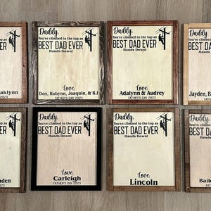 May include: Eight framed wood signs with a light brown background and black text. Each sign features a silhouette of a lineman climbing a utility pole. The text reads "Daddy, you've climbed to the top as BEST DAD EVER Hands Down!" Each sign has a different name and date at the bottom.