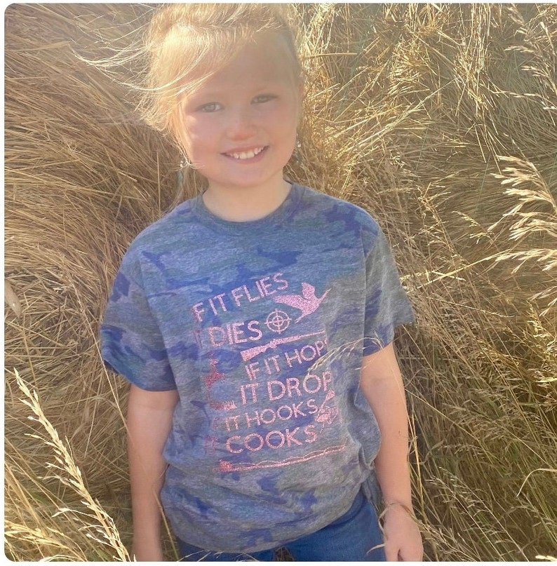 May include: A young person wearing a blue and green camouflage t-shirt with pink glitter text that reads "If it flies it dies, if it hops it drops, if it hooks it cooks".