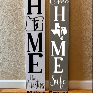 May include: Two wooden signs with the word "HOME" spelled vertically. The left sign is white with black lettering and a silhouette of a lineman working on a power line in the state of Oregon. The right sign is grey with white lettering and a silhouette of a lineman working on a power line in the state of Texas. The left sign says "The Martins" at the bottom and the right sign says "Come Home Safe".