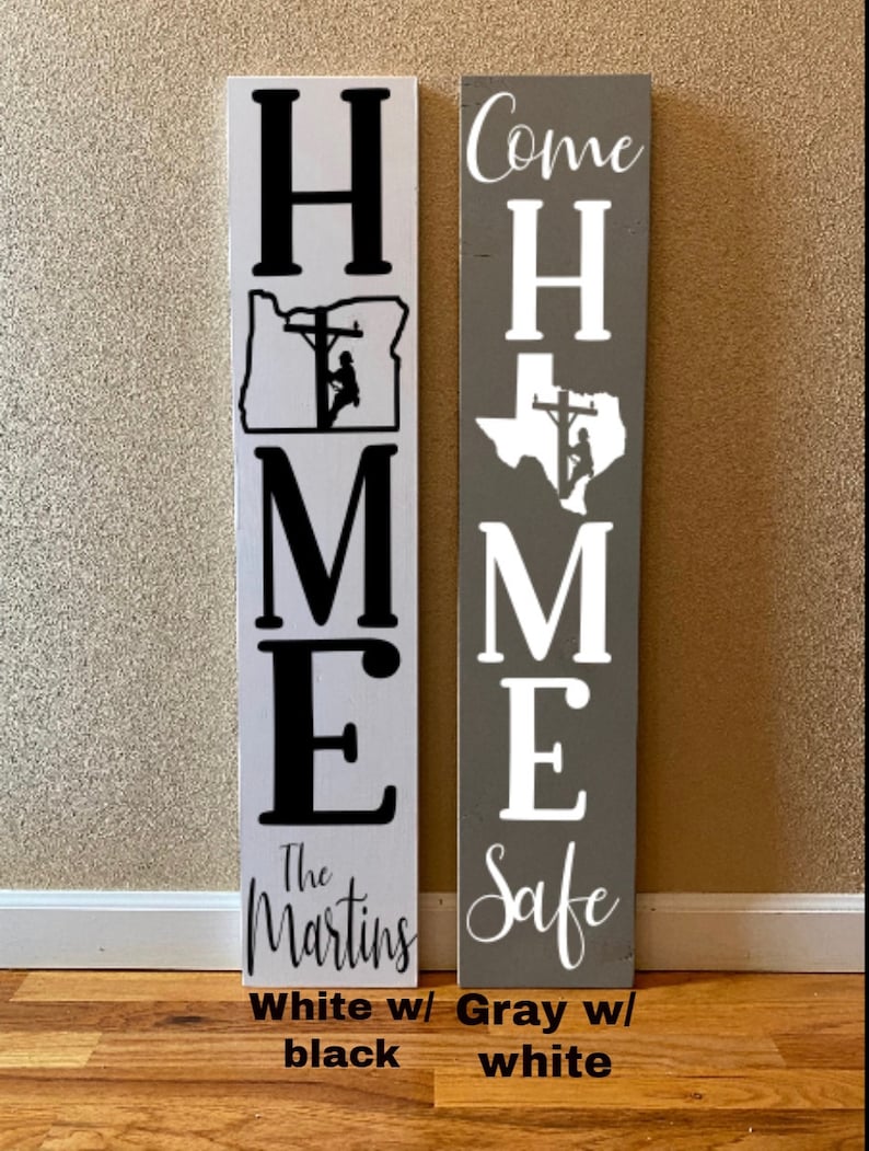 May include: Two wooden signs with the word "HOME" spelled vertically. The left sign is white with black lettering and a silhouette of a lineman working on a power line in the state of Oregon. The right sign is grey with white lettering and a silhouette of a lineman working on a power line in the state of Texas. The left sign says "The Martins" at the bottom and the right sign says "Come Home Safe".