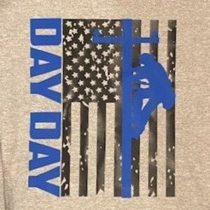 May include: A gray sweatshirt with a distressed American flag design. The flag is black and white with a blue silhouette of a lineman working on a power line. The words "Day Day" are printed vertically in blue on the left side of the flag.
