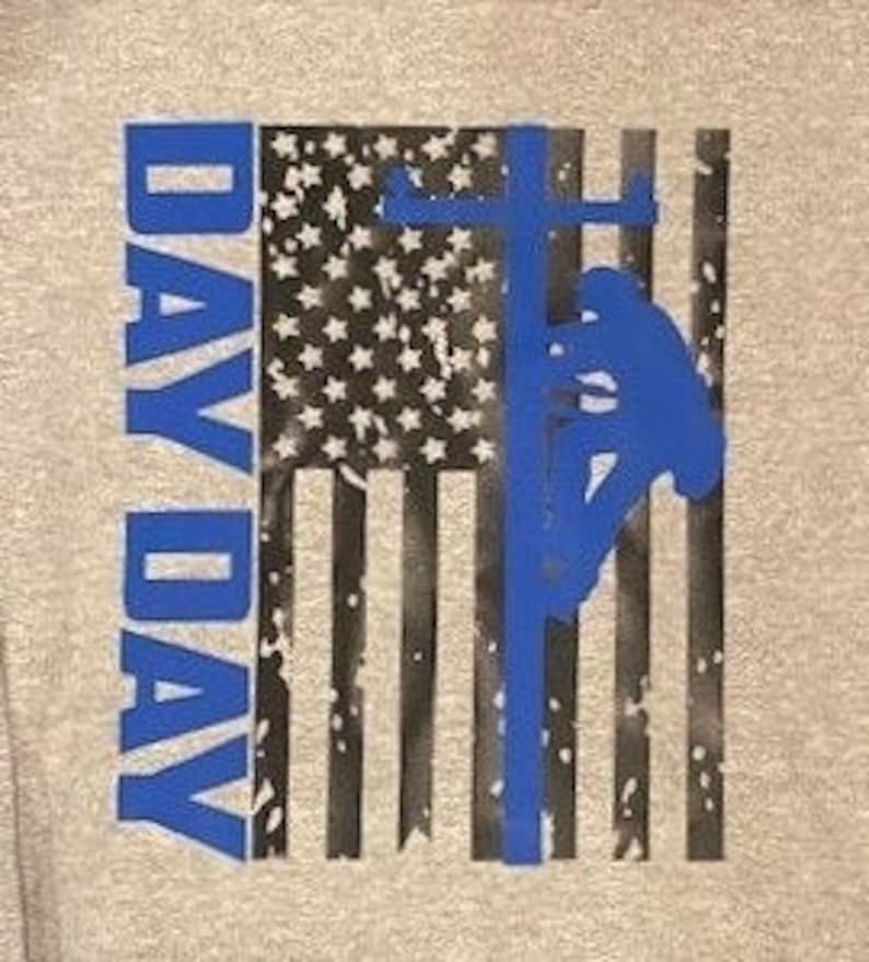 May include: A gray sweatshirt with a distressed American flag design. The flag is black and white with a blue silhouette of a lineman working on a power line. The words "Day Day" are printed vertically in blue on the left side of the flag.