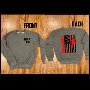 May include: A gray sweatshirt with the text "LINE KID" and a lightning bolt on the front. The back of the sweatshirt features a black and white American flag with a red silhouette of a lineman climbing a utility pole.