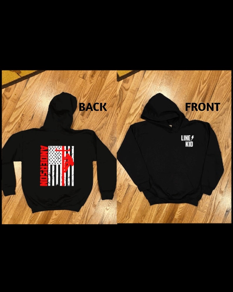 May include: Black hooded sweatshirt with a red, white, and blue American flag design on the back. The flag features a silhouette of a lineman climbing a utility pole. The text "Anderson" is printed above the flag. The front of the sweatshirt has the text "LINE KID" printed in white with a lightning bolt symbol.