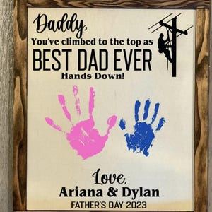 May include: A framed sign with a brown wood frame and a beige background. The sign says "Daddy, You've climbed to the top as BEST DAD EVER Hands Down!" with two handprints in pink and blue. The sign also says "Love, Ariana & Dylan FATHER'S DAY 2023".