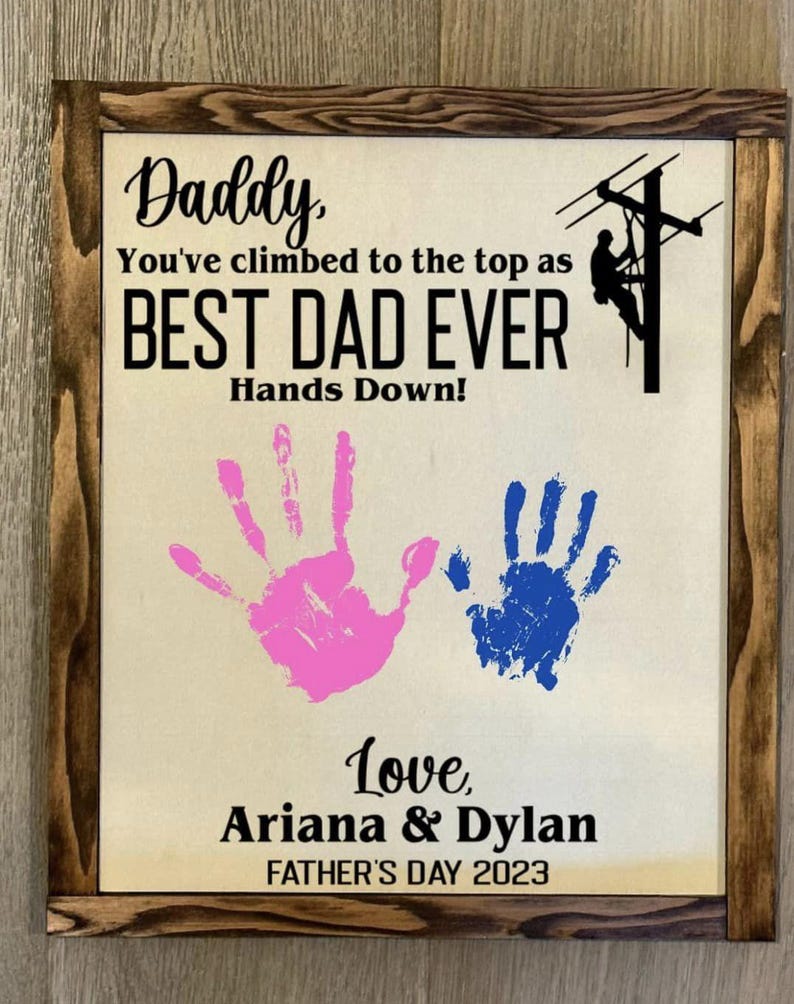 May include: A framed sign with a brown wood frame and a beige background. The sign says "Daddy, You've climbed to the top as BEST DAD EVER Hands Down!" with two handprints in pink and blue. The sign also says "Love, Ariana & Dylan FATHER'S DAY 2023".
