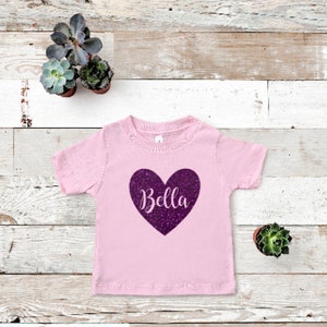 K&ouml;nnte beinhalten: Rosafarbenes T-Shirt mit kurzen &Auml;rmeln und einem glitzernden lilafarbenen Herz, auf dem "Bella" steht.