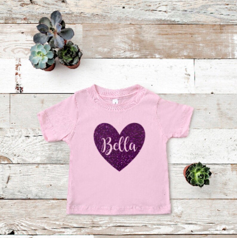 K&ouml;nnte beinhalten: Rosafarbenes T-Shirt mit kurzen &Auml;rmeln und einem glitzernden lilafarbenen Herz, auf dem "Bella" steht.