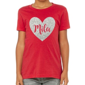 K&ouml;nnte beinhalten: Rotes T-Shirt f&uuml;r Kinder mit einem silbernen Glitzerherz, auf dem in roter Kursivschrift "Mila" steht.