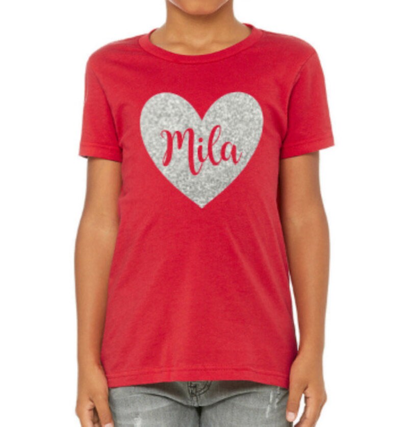 K&ouml;nnte beinhalten: Rotes T-Shirt f&uuml;r Kinder mit einem silbernen Glitzerherz, auf dem in roter Kursivschrift "Mila" steht.
