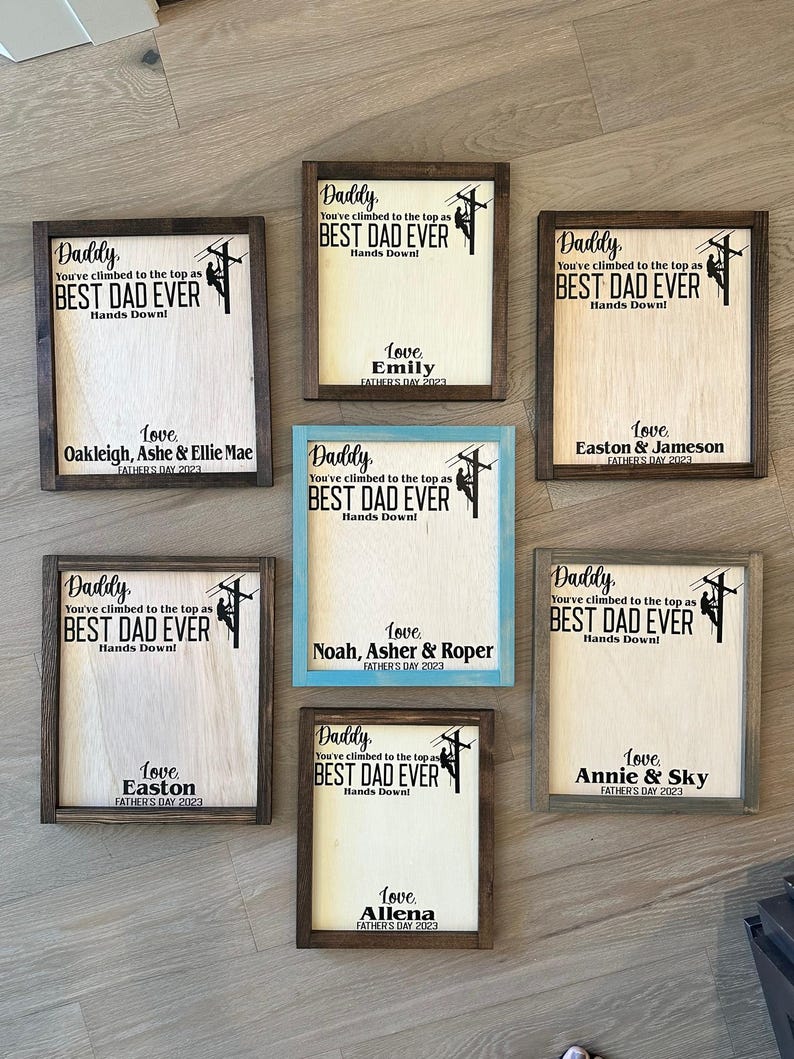 May include: Nine wooden signs with a brown frame and a light wood background. Each sign features a silhouette of a person climbing a utility pole with the text "Daddy, You've climbed to the top as BEST DAD EVER Hands Down!" and a personalized message with the name of the child or children and the year.