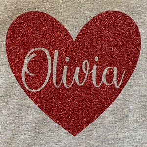 K&ouml;nnte beinhalten: Ein grauer Pullover mit einem roten Glitzerherz, auf dem "Olivia" in wei&szlig;em Glitzer steht.