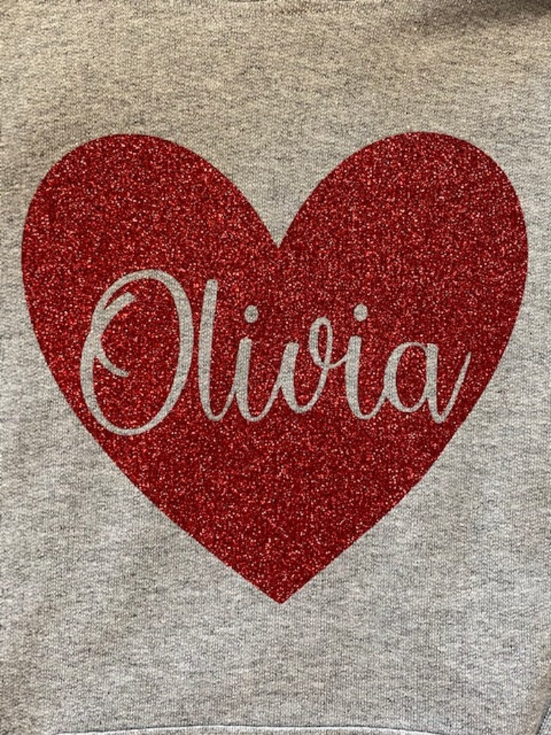 K&ouml;nnte beinhalten: Ein grauer Pullover mit einem roten Glitzerherz, auf dem "Olivia" in wei&szlig;em Glitzer steht.