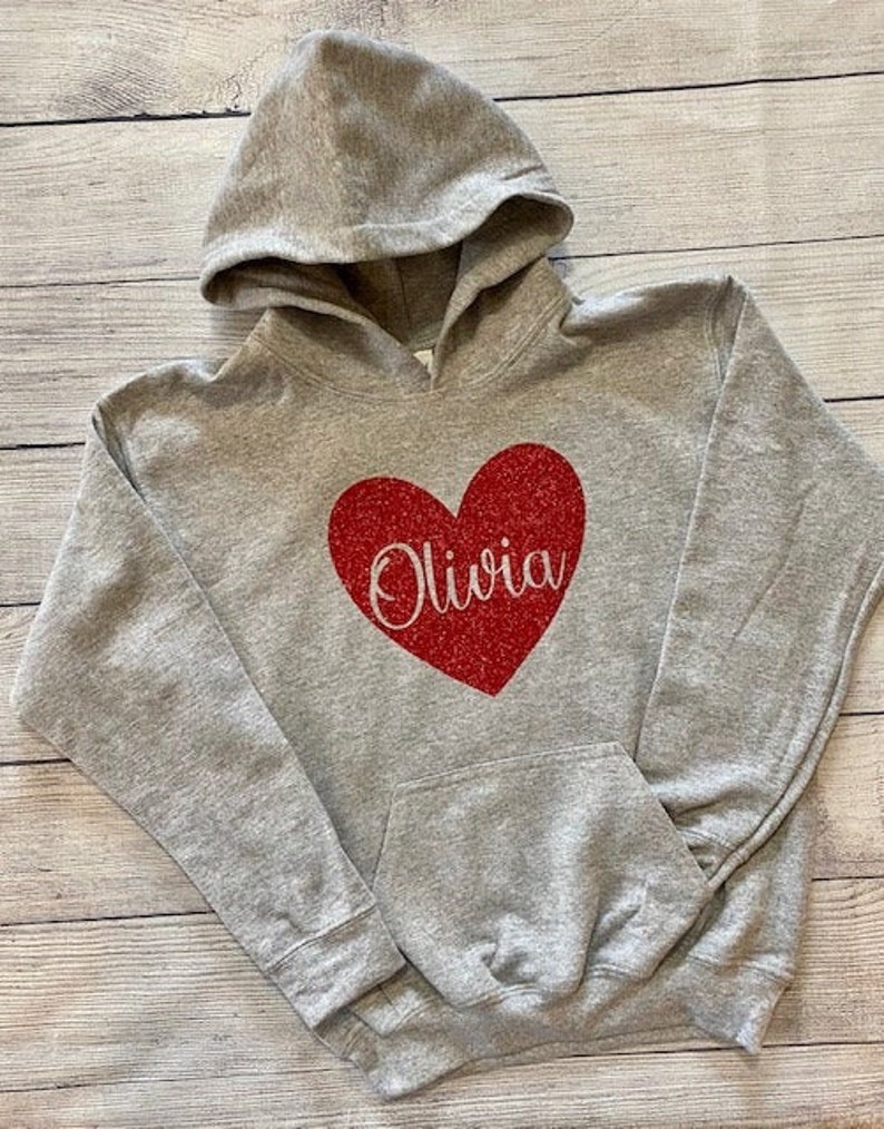 K&ouml;nnte beinhalten: Ein grauer Kapuzenpullover mit einem roten Glitzerherz und dem Namen "Olivia" in wei&szlig;em Glitzer.