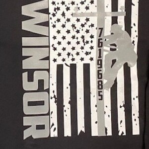 May include: Black and white graphic t-shirt with an American flag design. A silhouette of a person climbing a utility pole with the numbers 7619685 printed on the pole. The text "WINDSOR" is printed on the left side of the shirt.