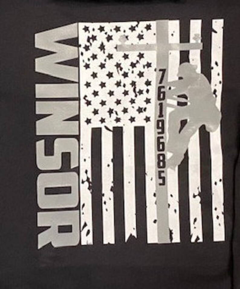 May include: Black and white graphic t-shirt with an American flag design. A silhouette of a person climbing a utility pole with the numbers 7619685 printed on the pole. The text "WINDSOR" is printed on the left side of the shirt.