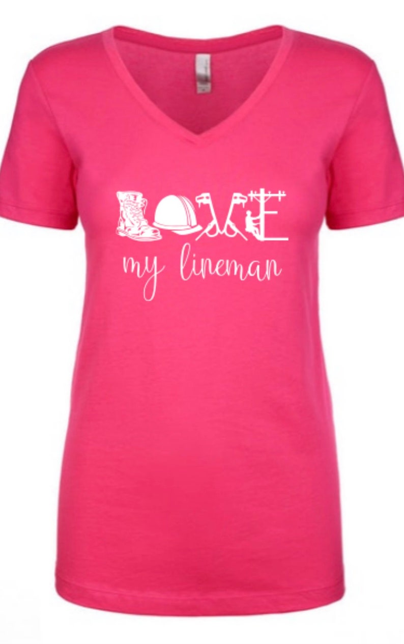 May include: Pink V-neck t-shirt with a white graphic design that says "Love my lineman" and includes illustrations of a hard hat, boots, and tools.