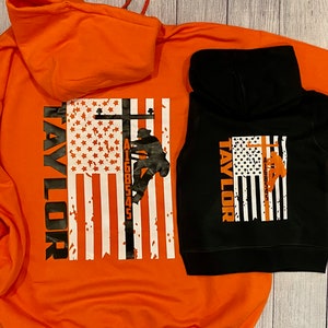 May include: Two hooded sweatshirts, one orange and one black, with a graphic of a lineman climbing a utility pole in front of an American flag. The text "TAYLOR" is printed vertically on the left side of the flag. The number "A1600545" is printed on the pole.